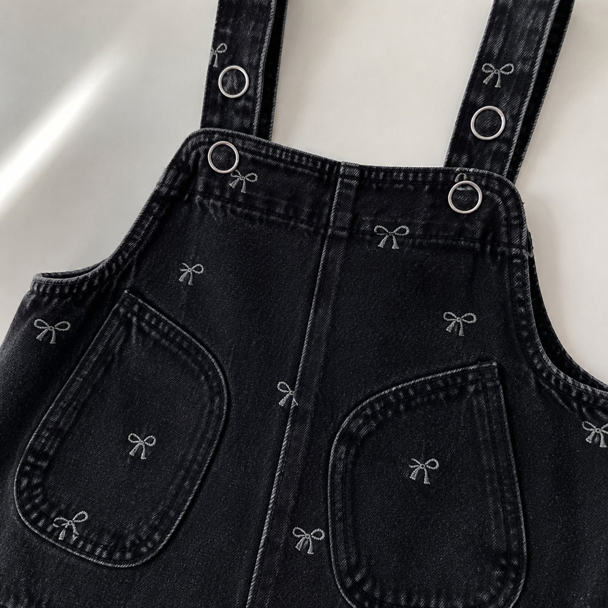Close-up of the black denim romper straps, hardware, pockets and bow motifs