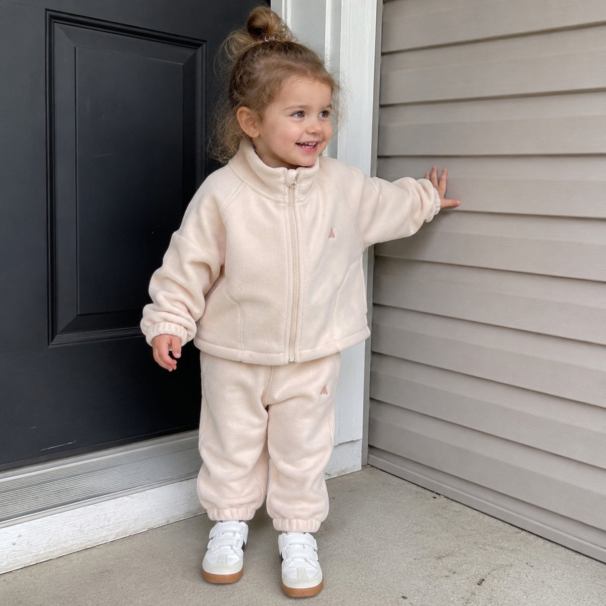 Child wearing beige fleece zip jacket and joggers near a home entryway