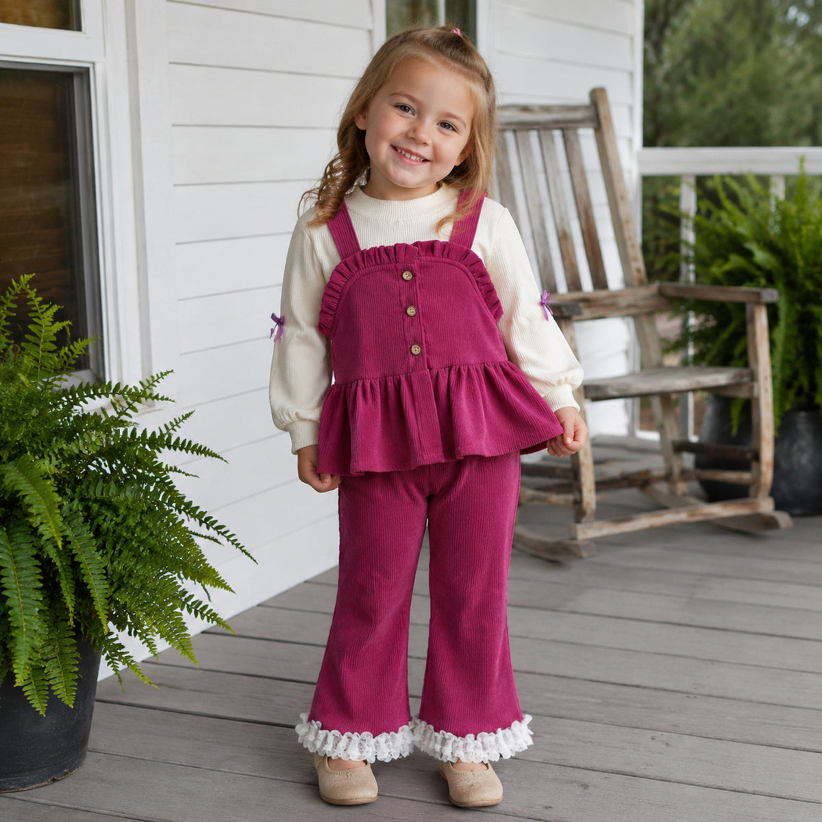 Girl wearing berry rose ruffle pinafore and pants set on a porch